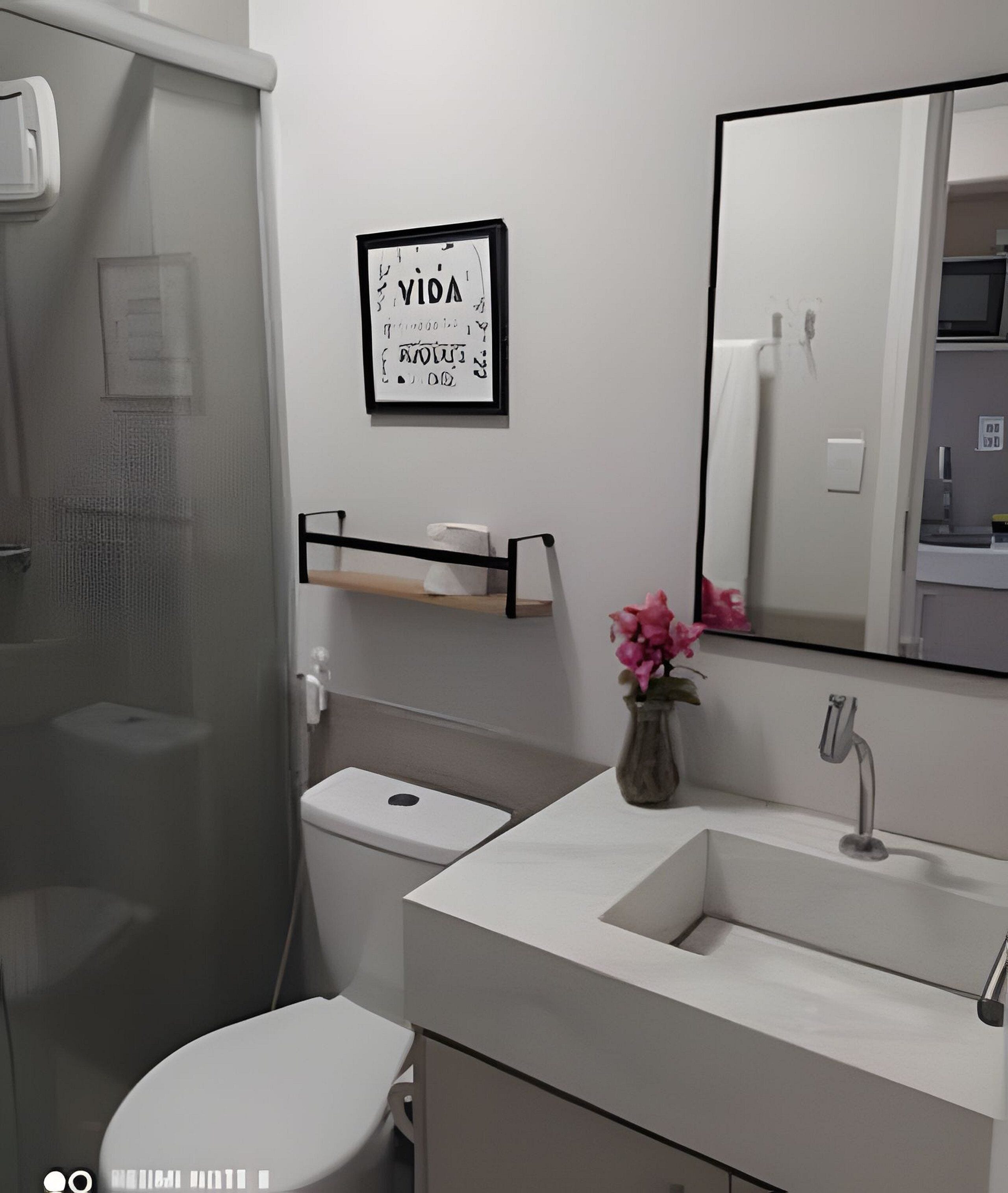 Uma casa de banho com sanita, lavatório e chuveiro. Há um letreiro emoldurado na parede e um vaso com flores no lavatório.