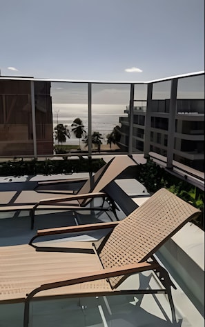 Uma área de piscina no terraço com espreguiçadeiras, vista para o oceano e edifícios modernos ao fundo.