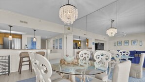 A modern kitchen with stainless steel appliances, a dining area with white chairs and a glass table, and a living room with a blue sofa and decorative wall art. - Oceanfront 3BR Condo w Pool, Balcony Views & Direct Beach Access (Wrightsville Beach)