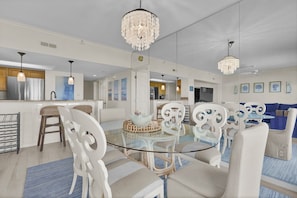 Eine moderne Küche mit Edelstahlgeräten, ein Essbereich mit weißen Stühlen und einem Glastisch sowie ein Wohnzimmer mit einem blauen Sofa und Wanddekoration.