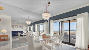 A modern dining area with a glass table, white chairs, and a chandelier. - Oceanfront 3BR Condo w Pool, Balcony Views & Direct Beach Access (Wrightsville Beach)