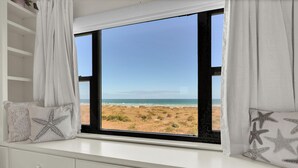 A room with a large window overlooking a beach, a white dresser, and star-patterned pillows. - Oceanfront 3BR Condo w Pool, Balcony Views & Direct Beach Access (Wrightsville Beach)