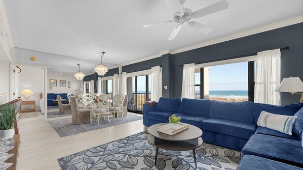 A modern living room with a blue sofa, a dining area with white chairs, and a view of the ocean. - Oceanfront 3BR Condo w Pool, Balcony Views & Direct Beach Access (Wrightsville Beach)