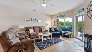 Una sala de estar con un sofá, sillones, una mesa de centro y un televisor. Hay un cartel que dice 'LAKE HOUSE' y una vista de un lago a través de la ventana.