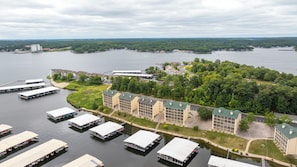 A riverside hotel with multiple docked boats, a large building with a green roof, and a forested area in the background. - WATERFRONT Condo/Bristol Bay/Screened-in balcony/Boat Slip Available & Garage (Village of Four Seasons)