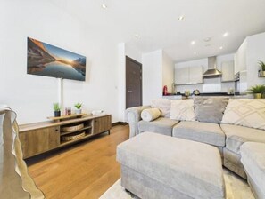 Una sala moderna con un sofá, un televisor de pantalla plana, un mueble de entretenimiento de madera y una cocina al fondo.