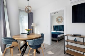 A modern room with a round wooden dining table, blue upholstered chairs, a lamp, a bed with a blue headboard, a TV, and a decorative mirror on the wall. - Wavescape - Wave Resort & Spa (Międzyzdroje)