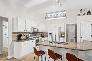 Eine Küche mit weißen Schränken, Granit-Arbeitsplatten, einer zentralen Kochinsel mit Barhocker, einer Spüle aus Edelstahl und einer hängenden Leuchte.