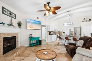 Ein geräumiges Wohnzimmer mit Kamin, Deckenventilator, Küchenbereich und Essplatz mit Barhocker.
