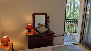 Une chambre avec une commode en bois, un miroir, une table de chevet avec une lampe, et une vue sur un balcon aménagé avec du mobilier d’extérieur.