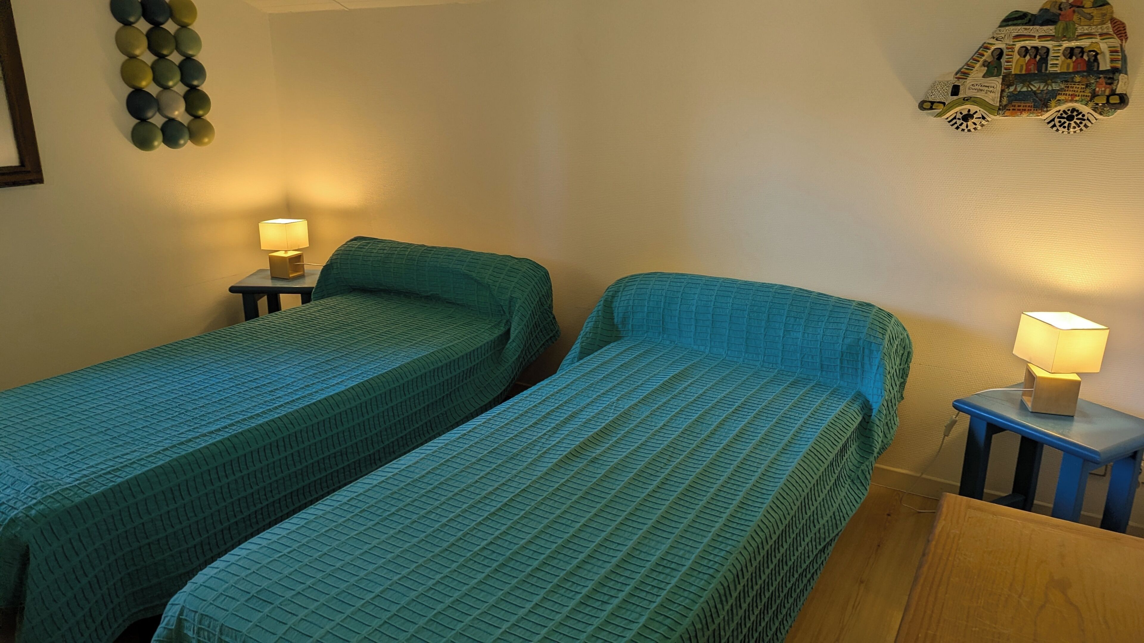 A room with two single beds, a small table, a nightstand with a lamp, and a wall decoration.