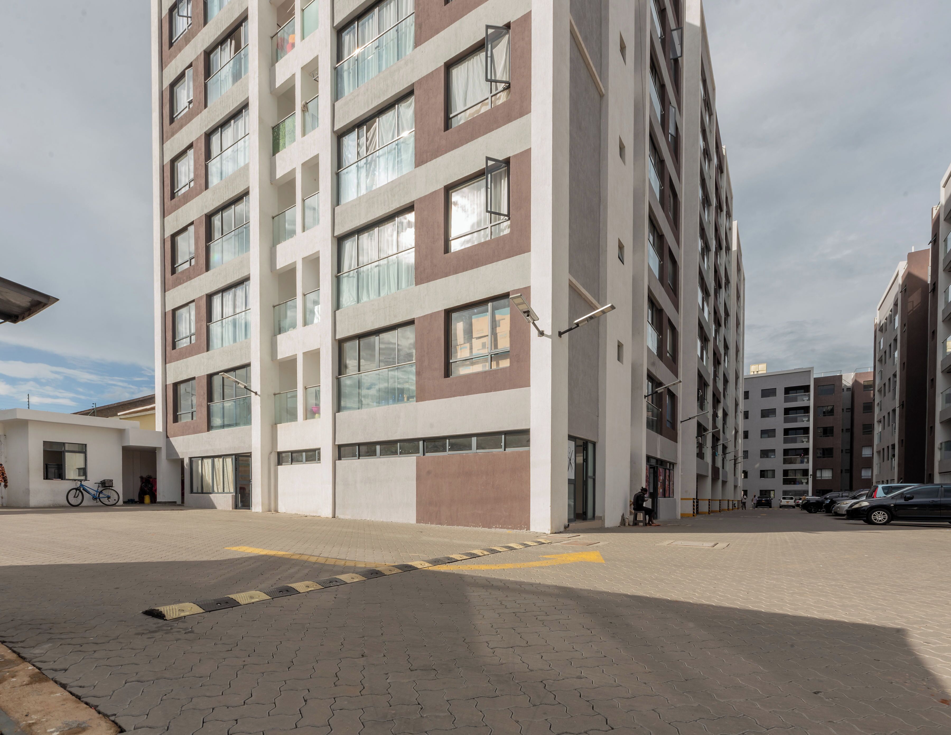 A multi-story residential building with a modern architectural design, featuring a mix of glass and concrete facades, and a paved ground with a yellow tactile paving strip.