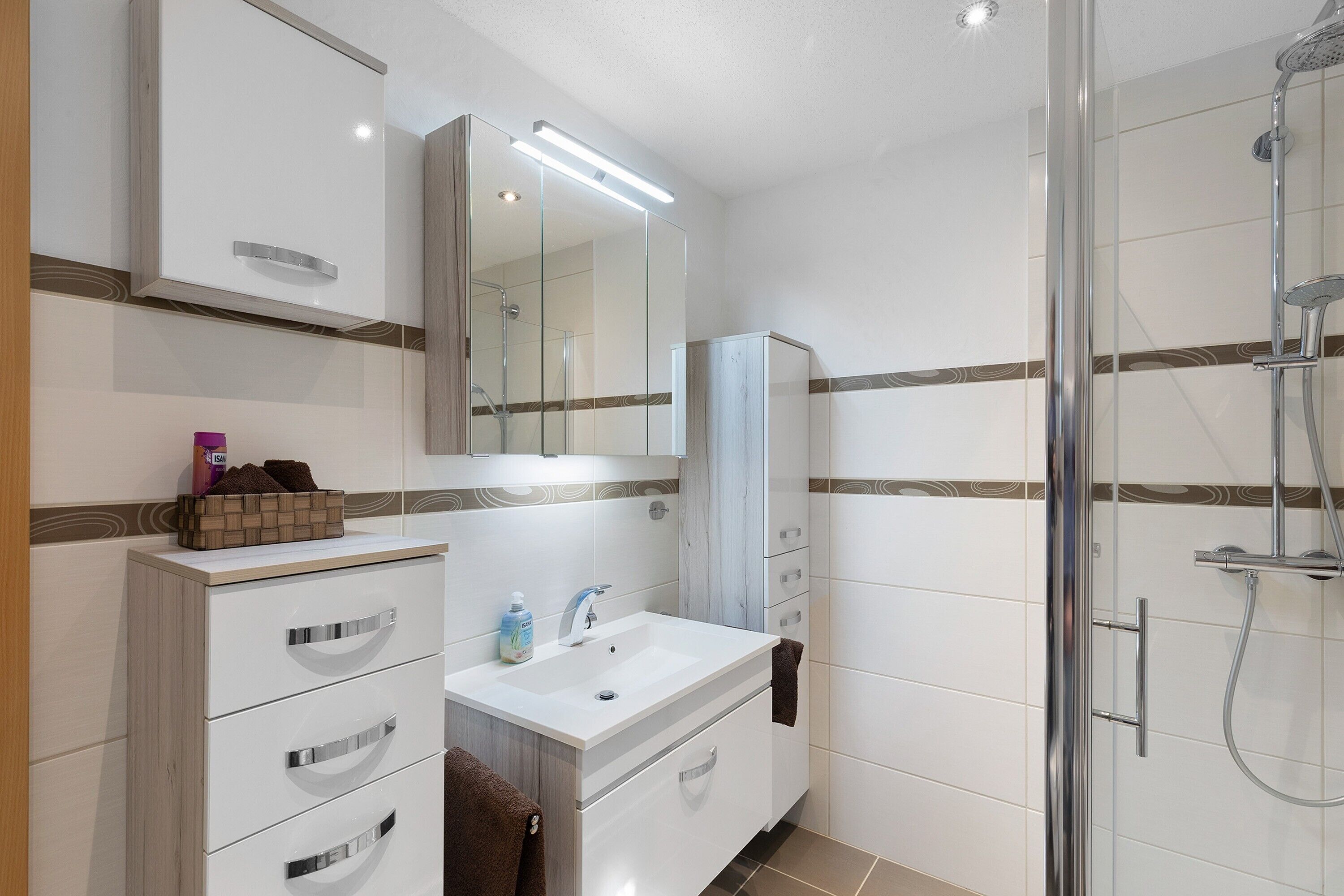 Een moderne badkamer met een douchecabine van glas, een witte wastafel met lades, een spiegel en een handdoekhouder.