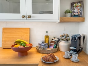 Una cocina con un frutero, un croissant, tazas de café, una cafetera y un libro de cocina en la estantería.