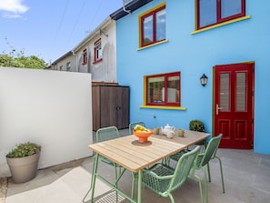 Un patio con una mesa de madera y sillas verdes, una puerta roja y una pared azul con ventanas.