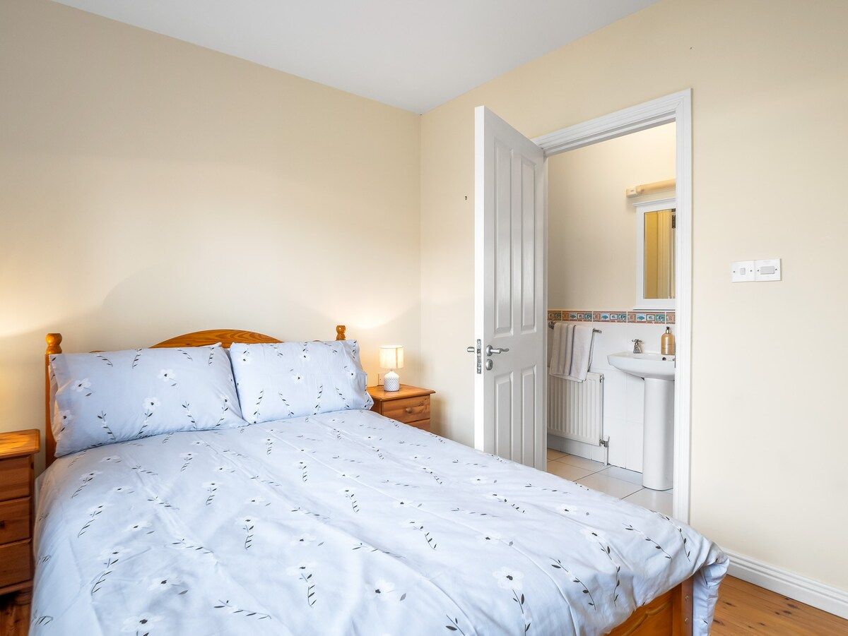 Quarto com cama, mesas de cabeceira, porta para um banheiro e piso ladrilhado.