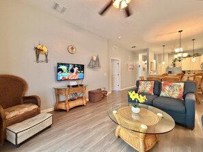 Ein Wohnzimmer mit Fernseher, Korbmöbeln, einem Glastisch und einer Couch mit floralen Kissen.