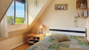 Un dormitorio con techo inclinado, una cama con colcha colorida, una lámpara de noche, una mesita de noche con un reloj y una ventana con vista a follaje.