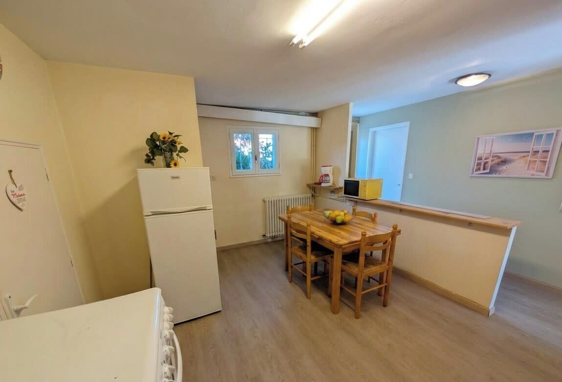 A compact kitchen with a refrigerator, a dining table with chairs, a window, and a radiator.