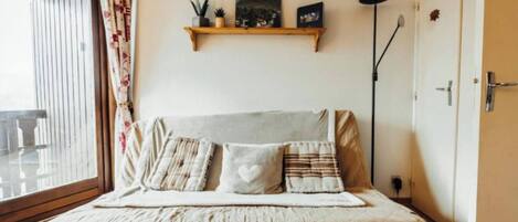 A cozy room with a bed, a window with curtains, a shelf with decorative items, and a door.