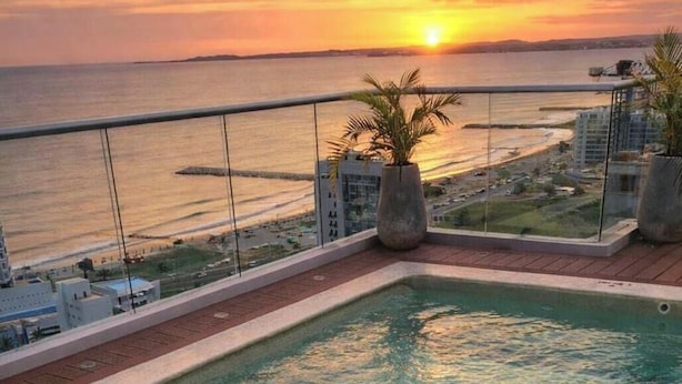 Une piscine aux eaux cristallines, avec vue sur le coucher de soleil sur l’océan, un palmier et un bâtiment moderne en arrière-plan.