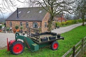 Ein grüner Traktor mit einer Ladefläche, ein Ziegelhaus mit Satteldach und ein Schotterweg.
