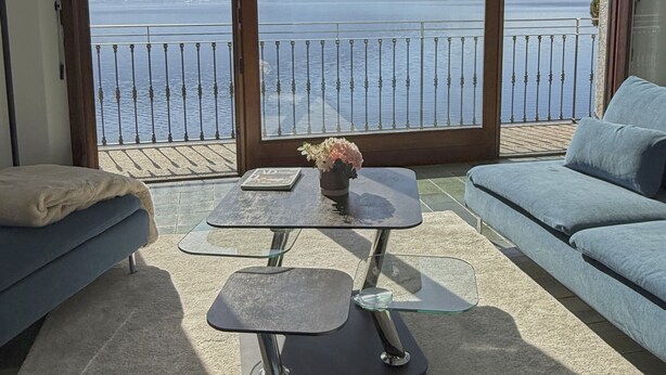 Un balcone con vista su un lago e montagne, un divano, un tavolo con un vaso e una lampada.