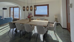 Zona pranzo con un grande tavolo in legno, sei sedie, lampade a sospensione e un vaso di fiori.
