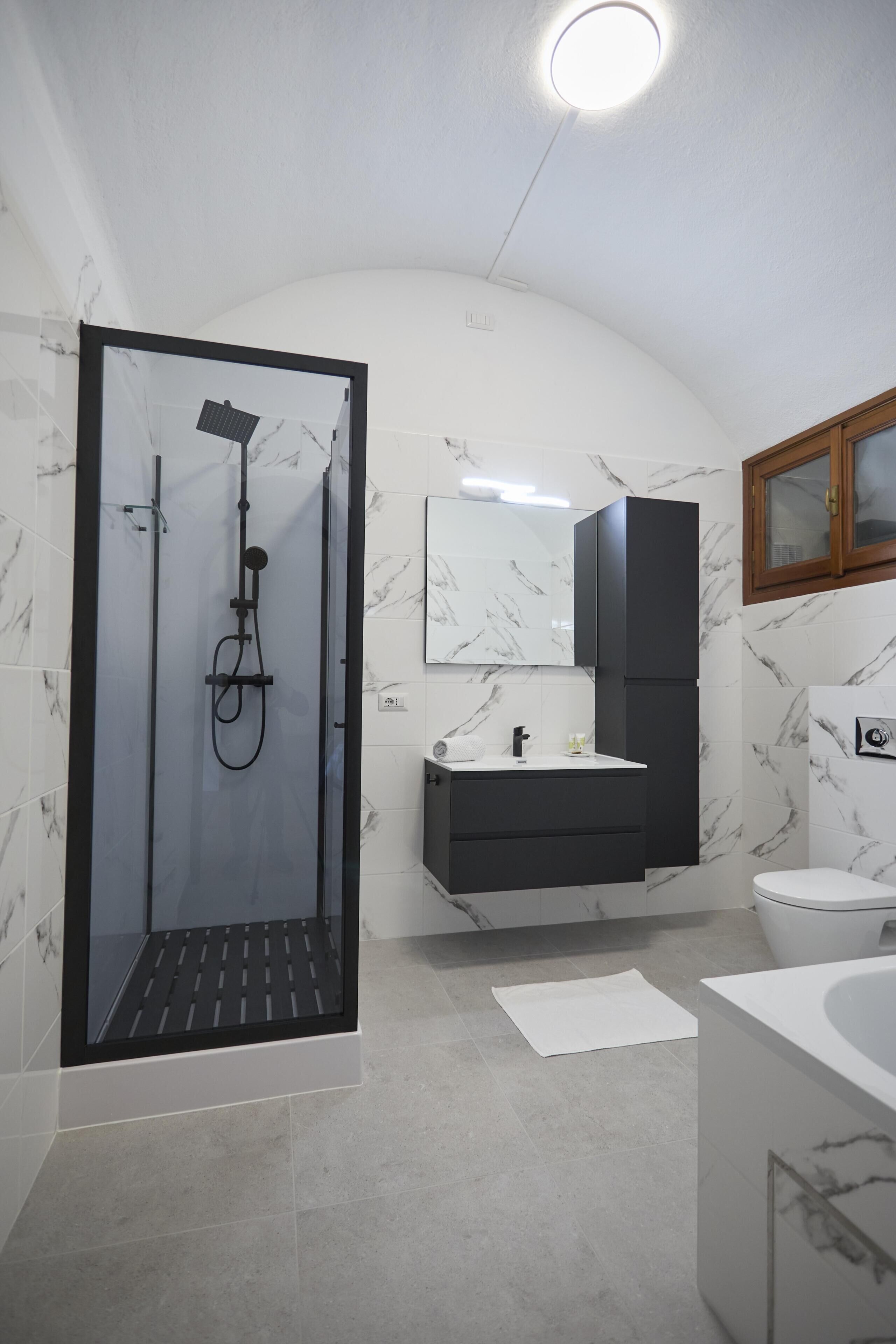 Un baño moderno con ducha envuelta en vidrio, un lavamanos blanco con mueble oscuro, un inodoro blanco y suelo de baldosas de mármol.