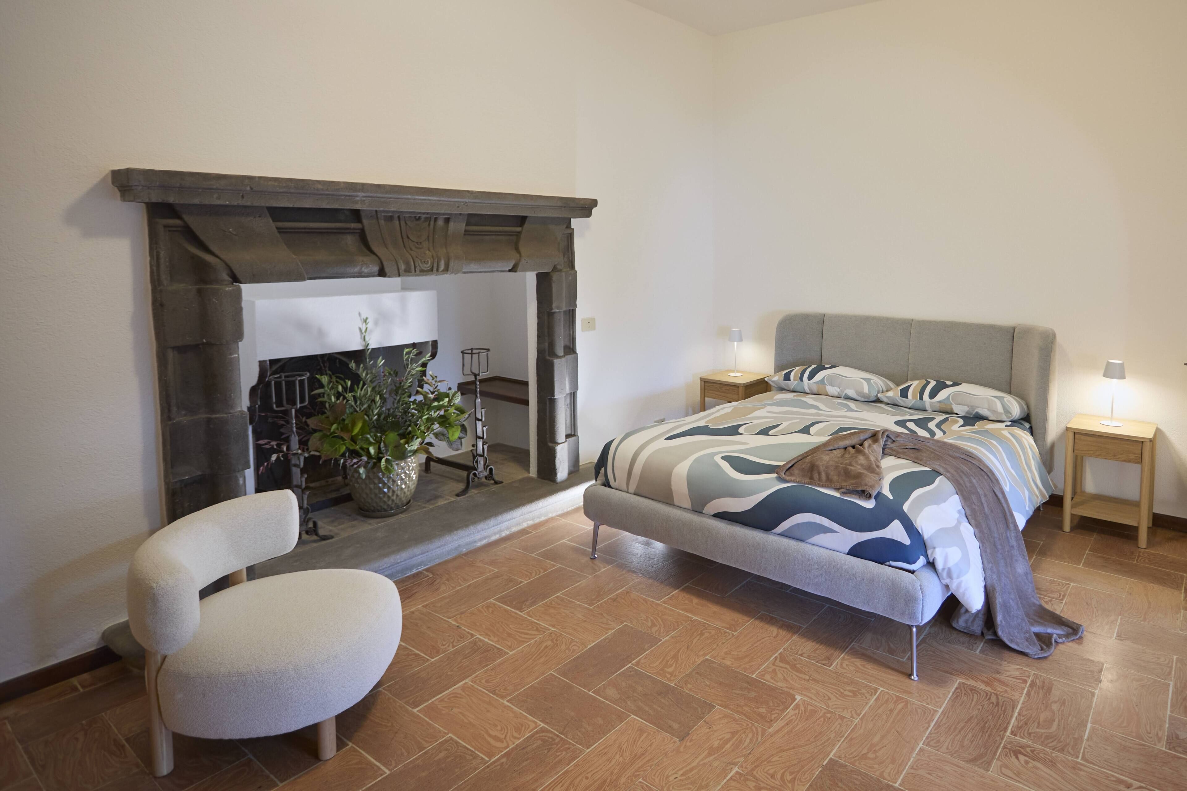 A bedroom with a bed, a chair, a fireplace, and a nightstand.