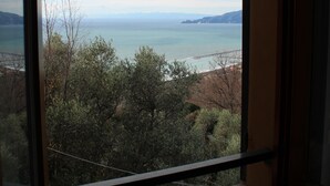 A room with a large window overlooking a coastal landscape, including trees and a distant shoreline. - A large terrace overlooking the sea in the quiet of olive trees for families (Santa Giulia)
