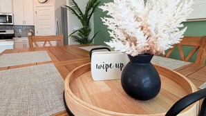 A dining area with a round wooden table, a black vase with white flowers, a 'wipe up' sign, and a green wall. - Your Perfect Getaway – Clean, Modern & Prime Location (Greer)