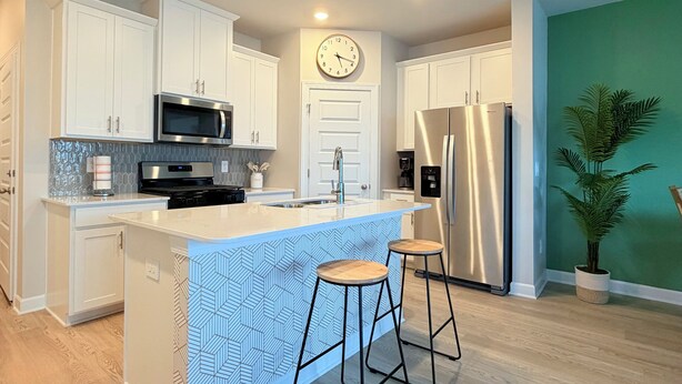 A modern kitchen with white cabinetry, a central island with stools, stainless steel appliances, and a clock on the wall. - Your Perfect Getaway – Clean, Modern & Prime Location (Greer)