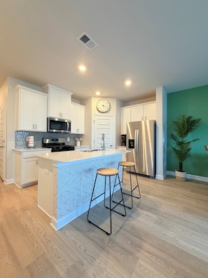 A modern kitchen with white cabinetry, a central island with stools, stainless steel appliances, and a clock on the wall.