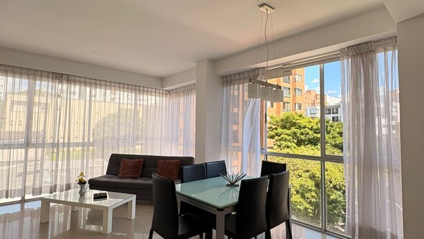 A spacious living area with a dining table, chairs, a sofa, and large windows offering a view of buildings and greenery.