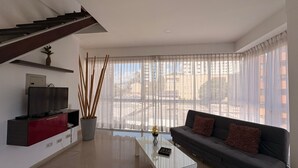 A modern living room with a staircase, a flat-screen TV, a sofa, a coffee table, and a large window with sheer curtains.