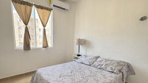 A bedroom with a bed, a window with curtains, an air conditioning unit, and a lamp on a nightstand. - Cancun apartment with pool, AC and Pet Friendly (Cancún)