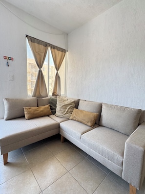 A living room with a beige sectional sofa, a window with curtains, and a wall-mounted light switch. - Cancun apartment with pool, AC and Pet Friendly (Cancún)