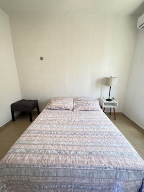 Ein Zimmer mit einem Einzelbett, einer Bettdecke mit Muster, einem Nachttisch mit Lampe und einem kleinen Hocker.