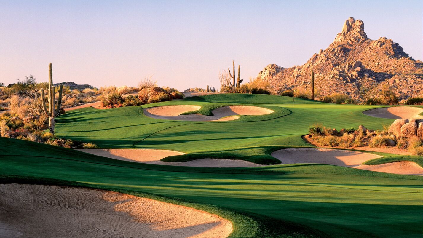 A desert golf course with sand bunkers, a cactus, and a rocky mountain in the background.