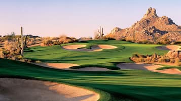 A desert golf course with sand bunkers, a cactus, and a rocky mountain in the background.