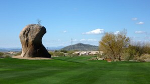 Un parcours de golf avec une formation rocheuse remarquable, un ciel dégagé et des montagnes à l’horizon.