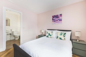 Une chambre à coucher avec un lit, deux tables de chevet avec des lampes, une œuvre d’art accrochée au mur, et une salle de bain visible à travers une porte ouverte.
