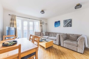 Un salon moderne avec une table à manger en bois, un canapé gris et une table basse en verre. Il y a deux luminaires au plafond et une grande fenêtre avec des rideaux.