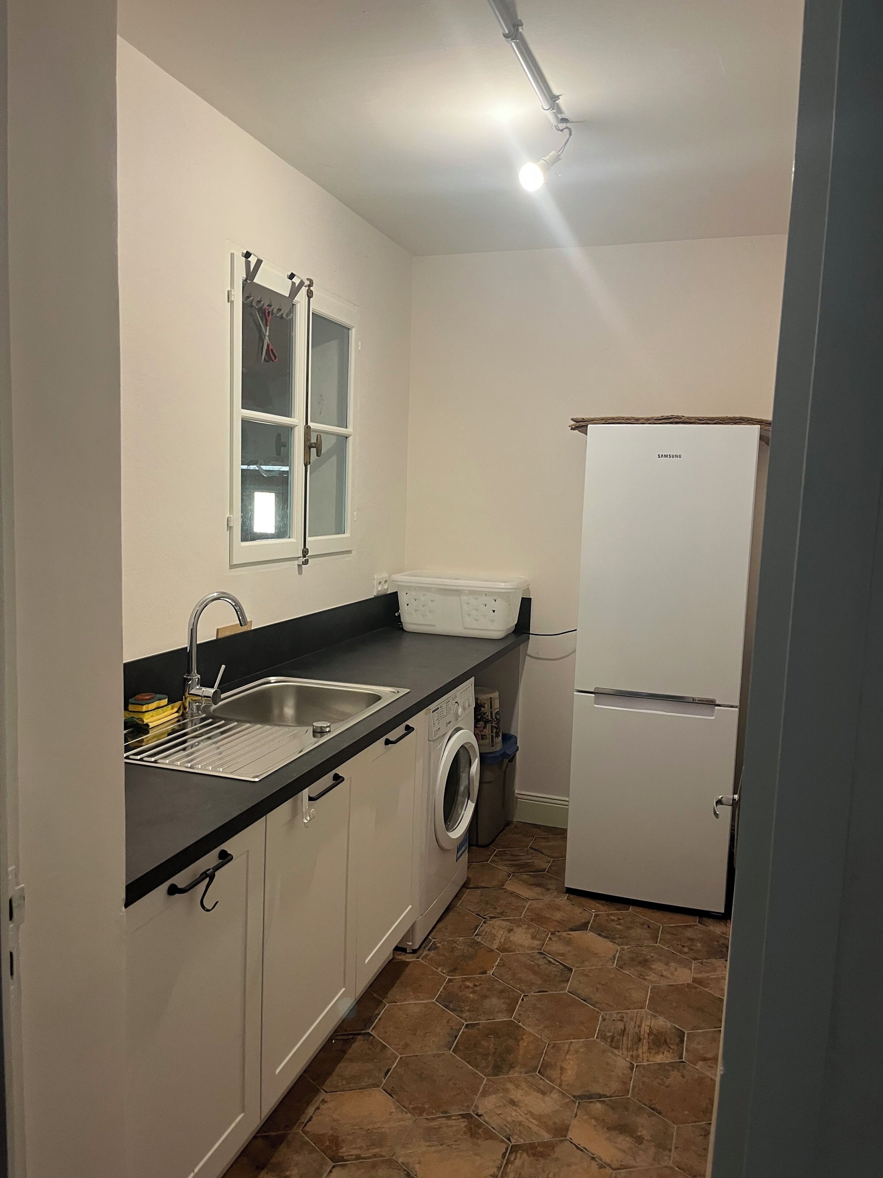 A compact kitchen with a sink, a washing machine, a refrigerator, and a window with blinds.