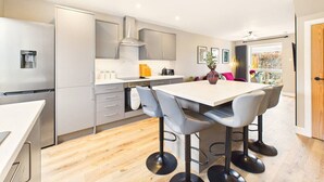 Una cocina moderna con gabinetes grises, una campana extractora de acero inoxidable y una zona de comedor con una mesa blanca y sillas grises.