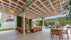 A covered outdoor patio with wooden furniture, a pool, and greenery. - 355 Luxury House | Patio | GameRoom | Pool | Grill (Davenport)