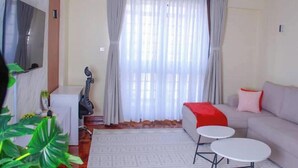 Ein Wohnzimmer mit einer Couch, zwei kleinen Tischen, einem Fernseher und einem großen Fenster mit Vorhängen.