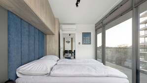 A modern bedroom with a large bed, a nightstand, a desk, and a view of the city through large windows. - Serenity - Wave Resort & Spa (Międzyzdroje)