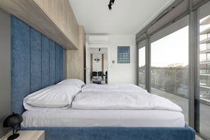 Ein modernes Schlafzimmer mit einem großen Bett, einem Nachttisch, einem Schreibtisch und Blick auf die Stadt durch große Fenster.